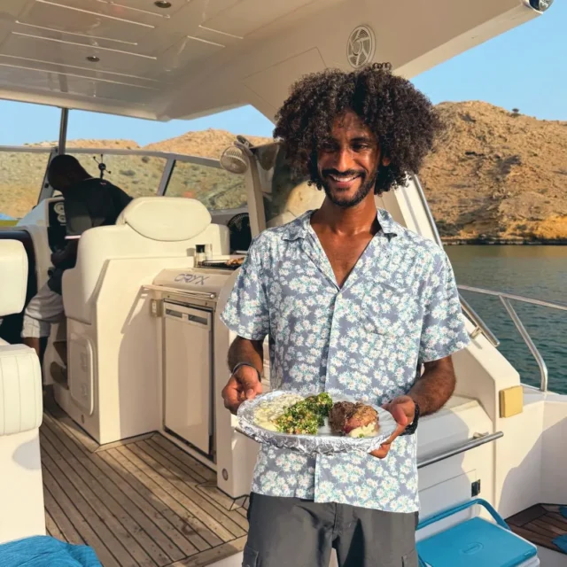 A smiling man on the yacht deck holding a plate of grilled meat, tabbouleh and dips